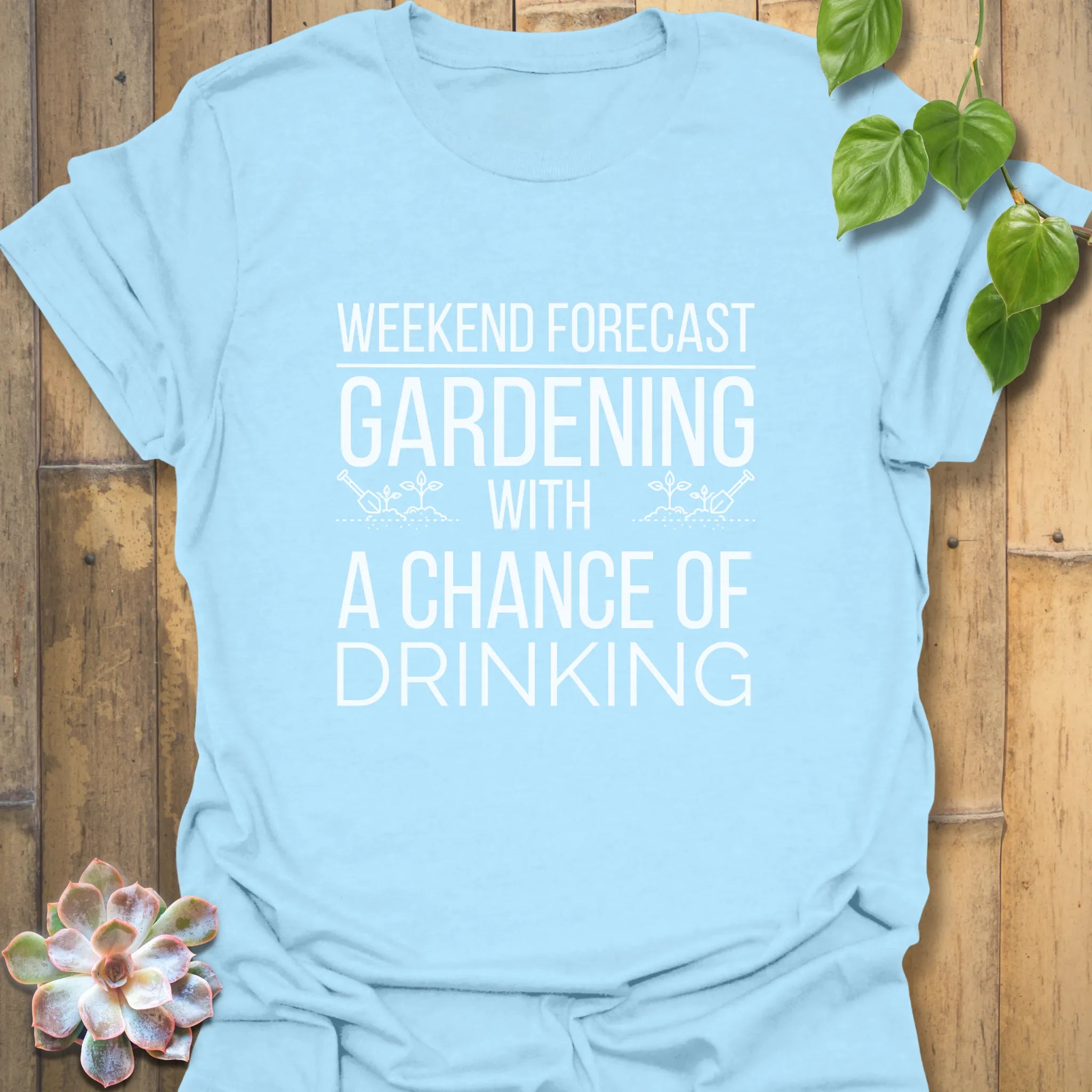 Light blue unisex T-shirt featuring the phrase “Gardening solves most of my problems, wine solves the rest” in white print, styled flat on a rustic wood surface with a green plant accent.