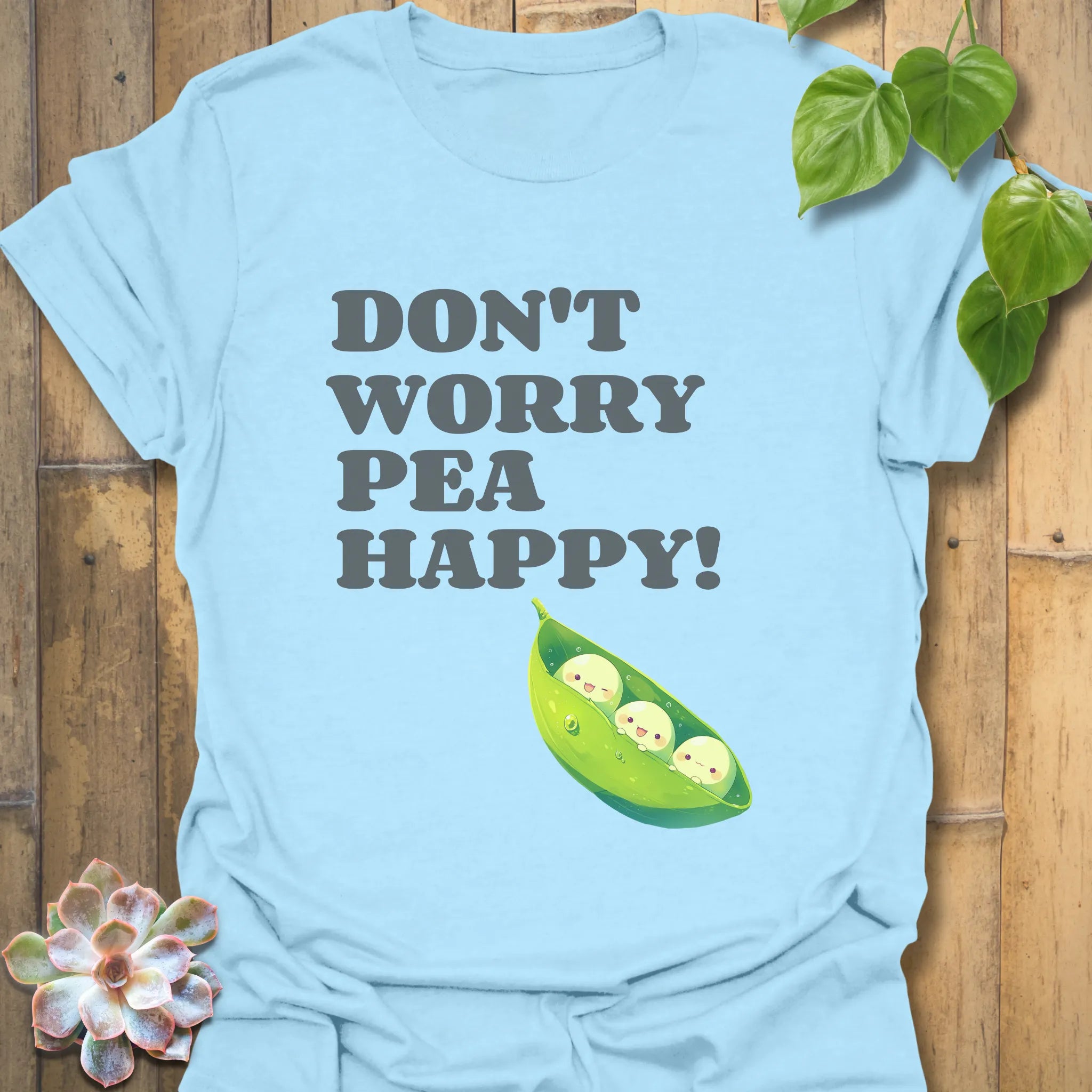 Light blue t-shirt featuring the design “Don’t Worry Pea Happy” in bold white letters with three smiling peas in a pod illustration