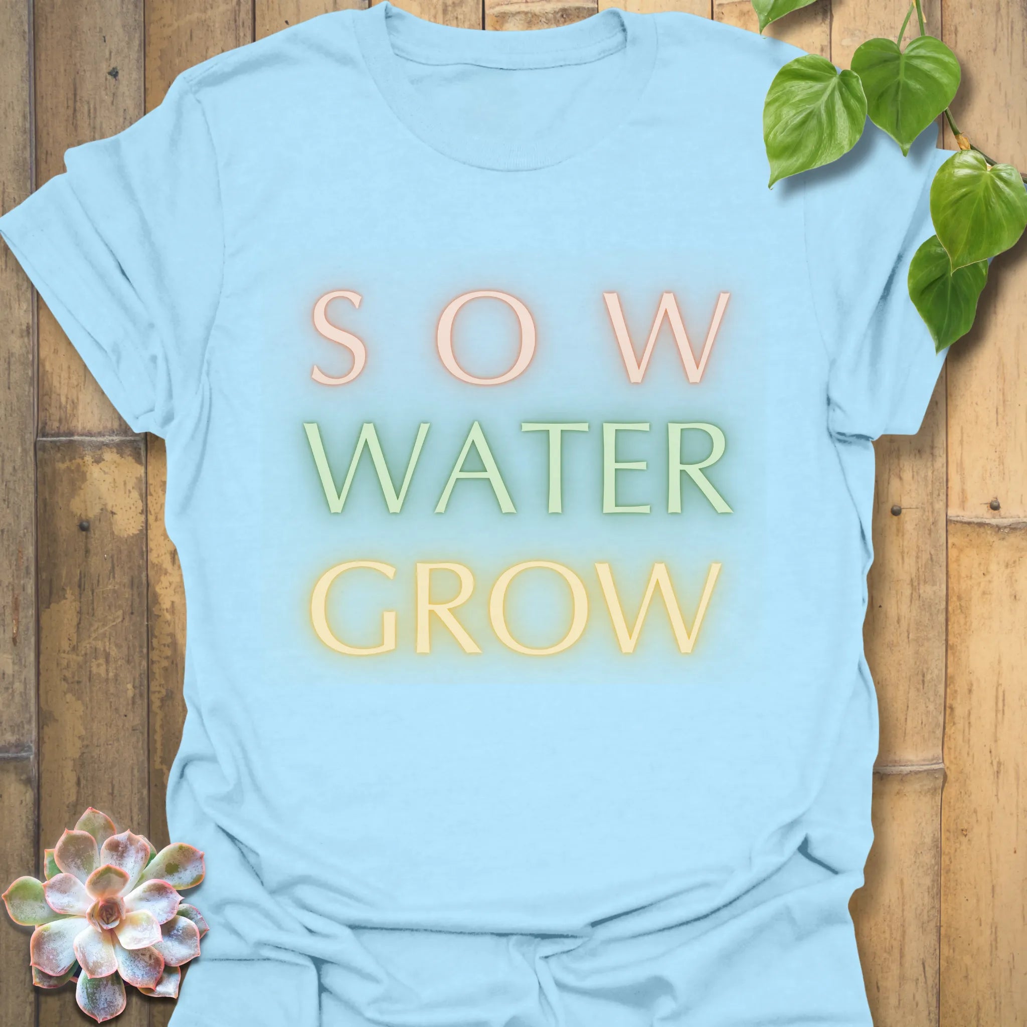 Light blue t-shirt featuring the design Sow Water Grow printed with a botanical watering can illustration.