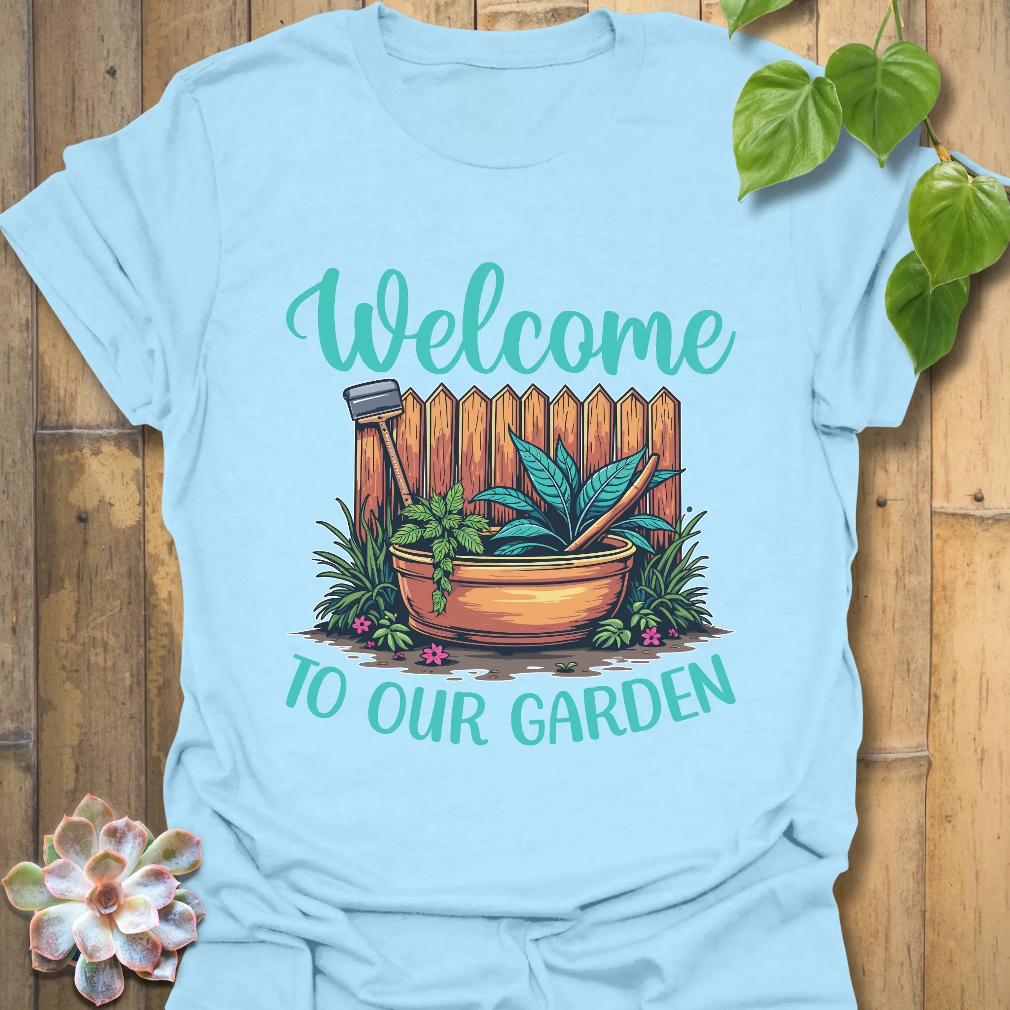 Light blue t-shirt featuring the design Welcome to our garden with a garden scene, potted plant, and tools, a fun garden tools graphic tee for plant enthusiasts.