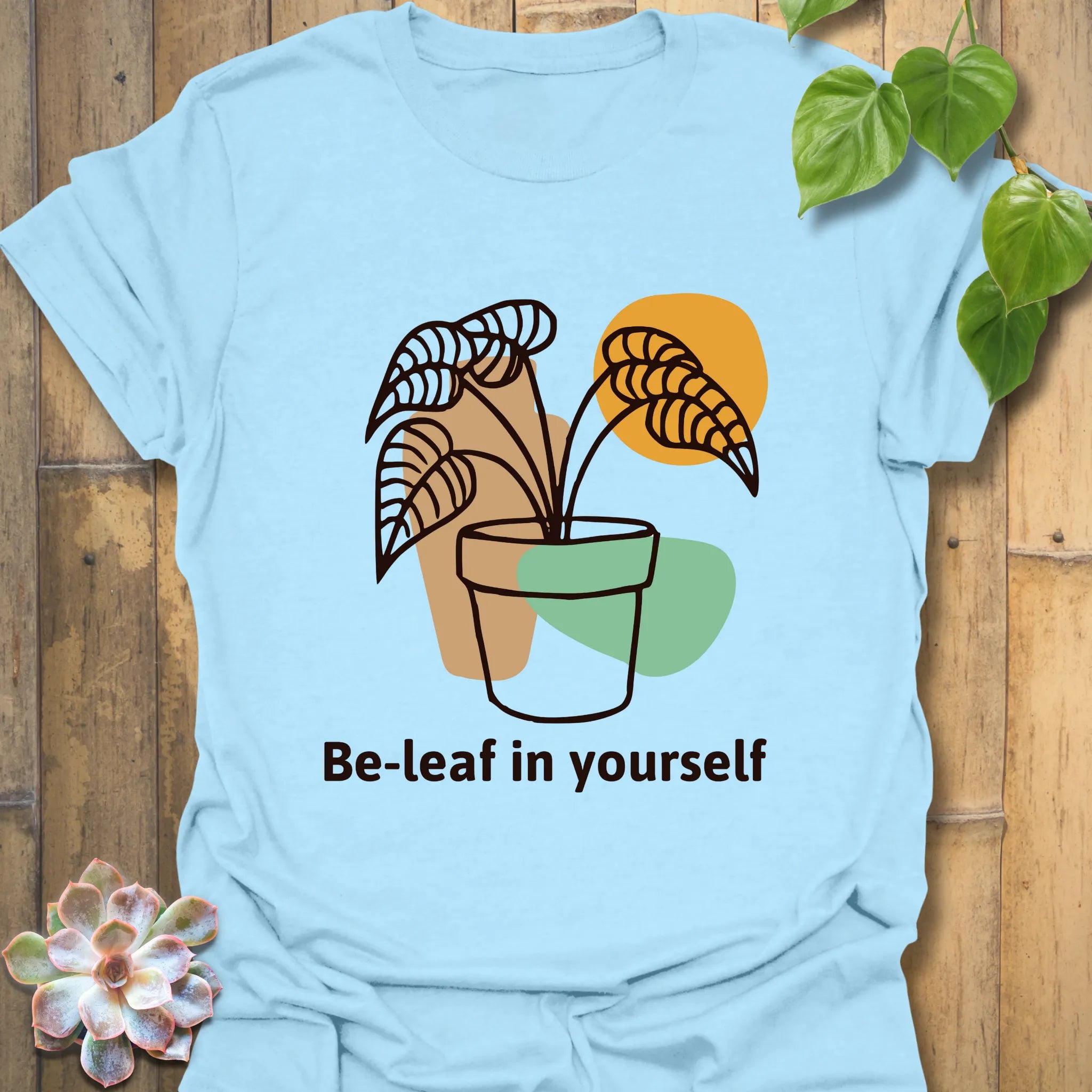 Light blue t-shirt with the “Be-leaf in yourself” slogan and a potted plant artwork