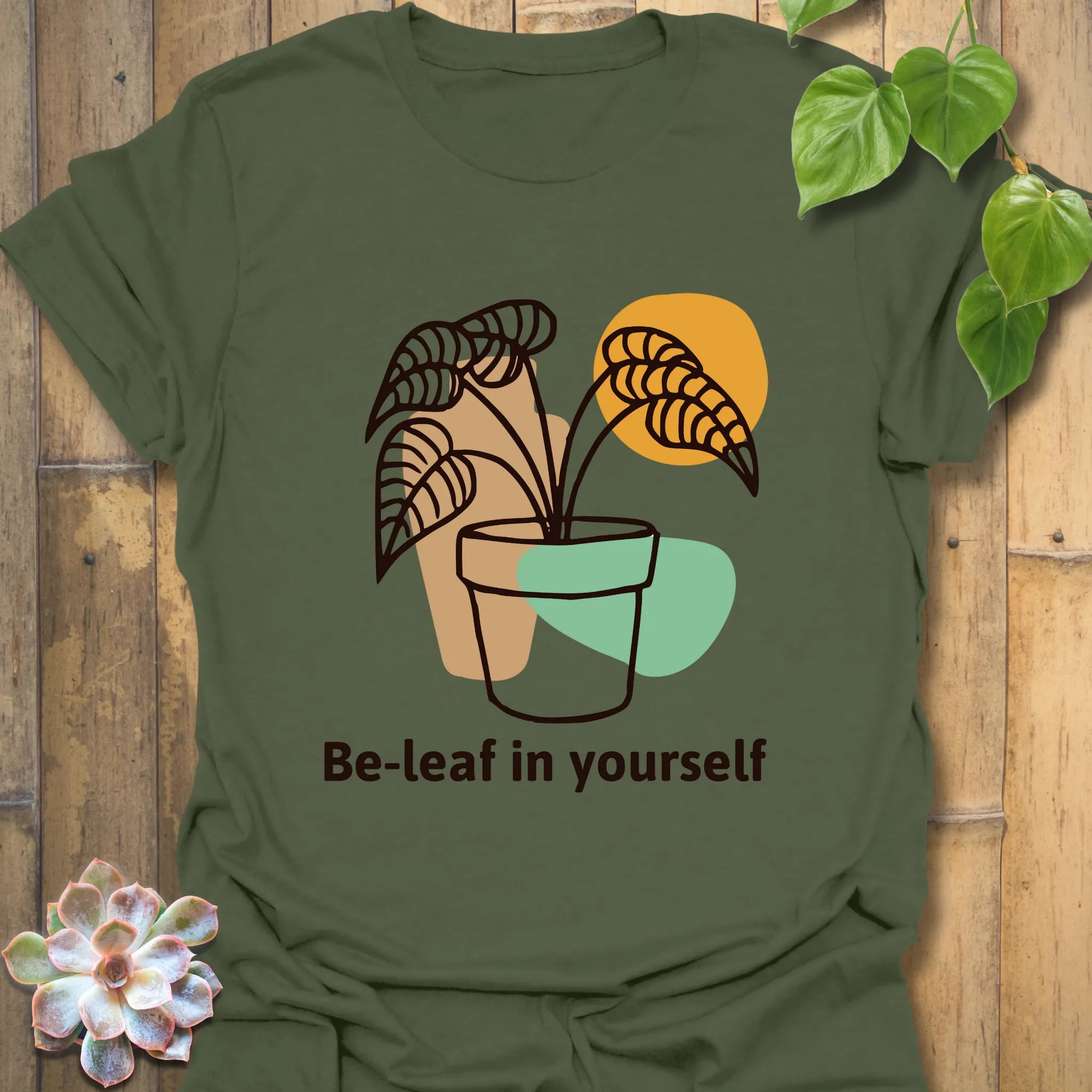Military green t-shirt featuring the phrase “Be-leaf in yourself” with a potted plant graphic