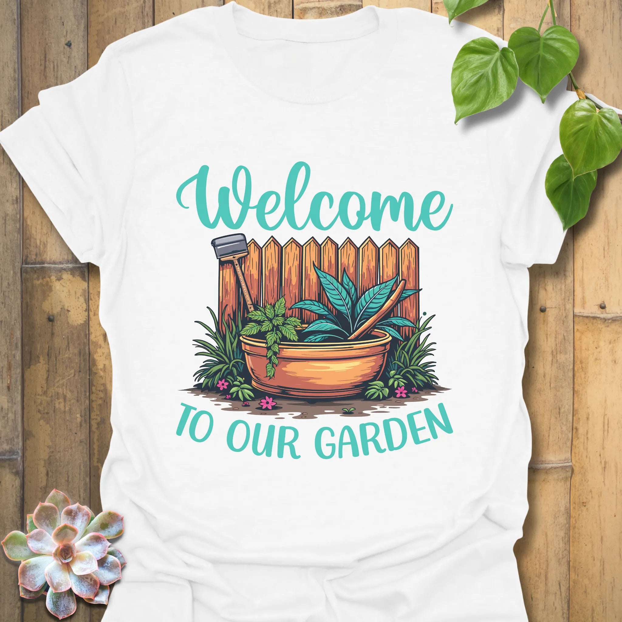 White t-shirt featuring the design Welcome to our garden with a garden scene, potted plant, and tools, perfect as a gardening graphic tee for plant lovers.