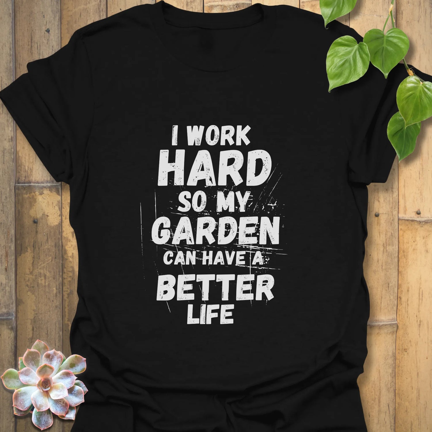 Black cotton T-shirt with bold white distressed print reading “I work hard so my garden can have a better life,” styled flat against a wooden backdrop with a succulent decoration.