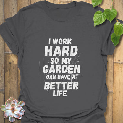Charcoal-coloured cotton T-shirt featuring bold white worn-style lettering that says “I work hard so my garden can have a better life,” laid flat with rustic wood and greenery accents.