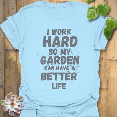 Light blue unisex T-shirt with bold white distressed text reading “I work hard so my garden can have a better life,” displayed flat on a wooden background with a succulent plant beside it.