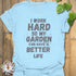 Light blue unisex T-shirt with bold white distressed text reading “I work hard so my garden can have a better life,” displayed flat on a wooden background with a succulent plant beside it.