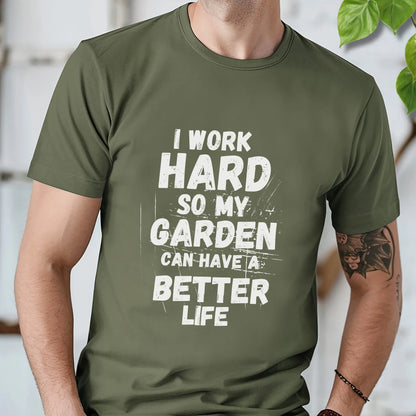 Military green unisex T-shirt with large white distressed text stating “I work hard so my garden can have a better life,” displayed flat on a timber background with plant leaves.