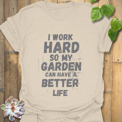 Natural cream-coloured unisex T-shirt featuring bold black distressed print that says “I work hard so my garden can have a better life,” laid flat on a rustic wood surface with plant accents.