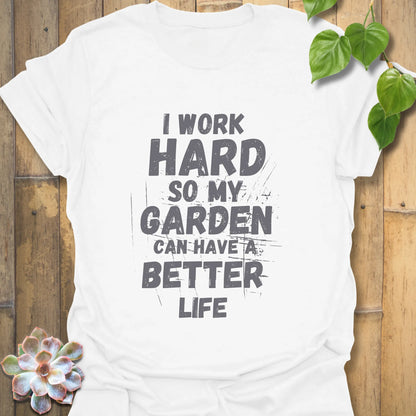 White cotton T-shirt with bold black distressed text reading “I work hard so my garden can have a better life,” displayed flat on a wooden background with greenery in the corner.

Natural