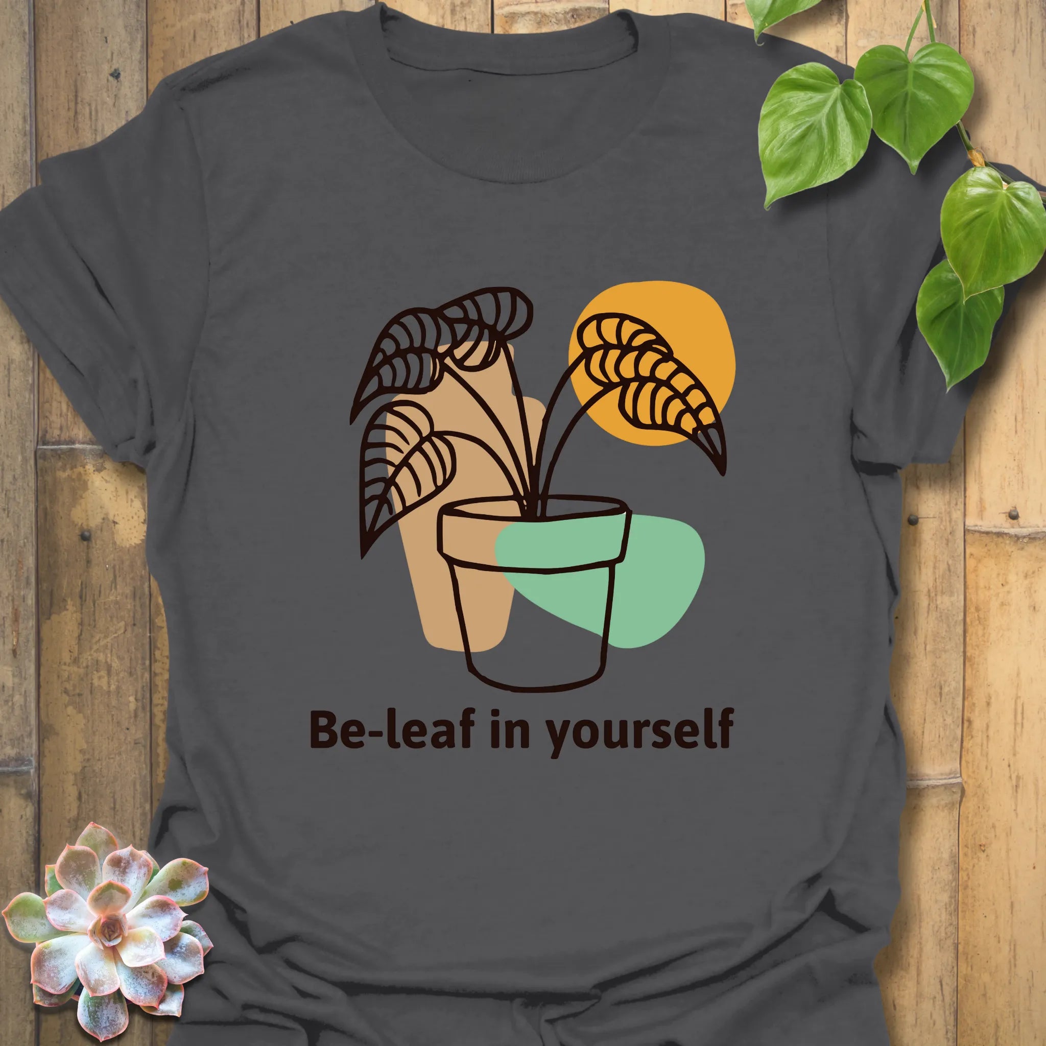 Charcoal grey t-shirt with the design “Be-leaf in yourself” showing a potted plant illustration on the front