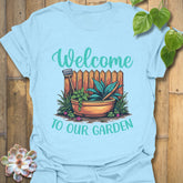 Light blue t-shirt featuring the design Welcome to our garden with a garden scene, potted plant, and tools, a fun garden tools graphic tee for plant enthusiasts.