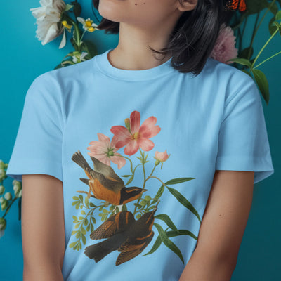 Light blue t-shirt with the Spring Songbirds illustration of two birds surrounded by soft pink flowers and leaves.