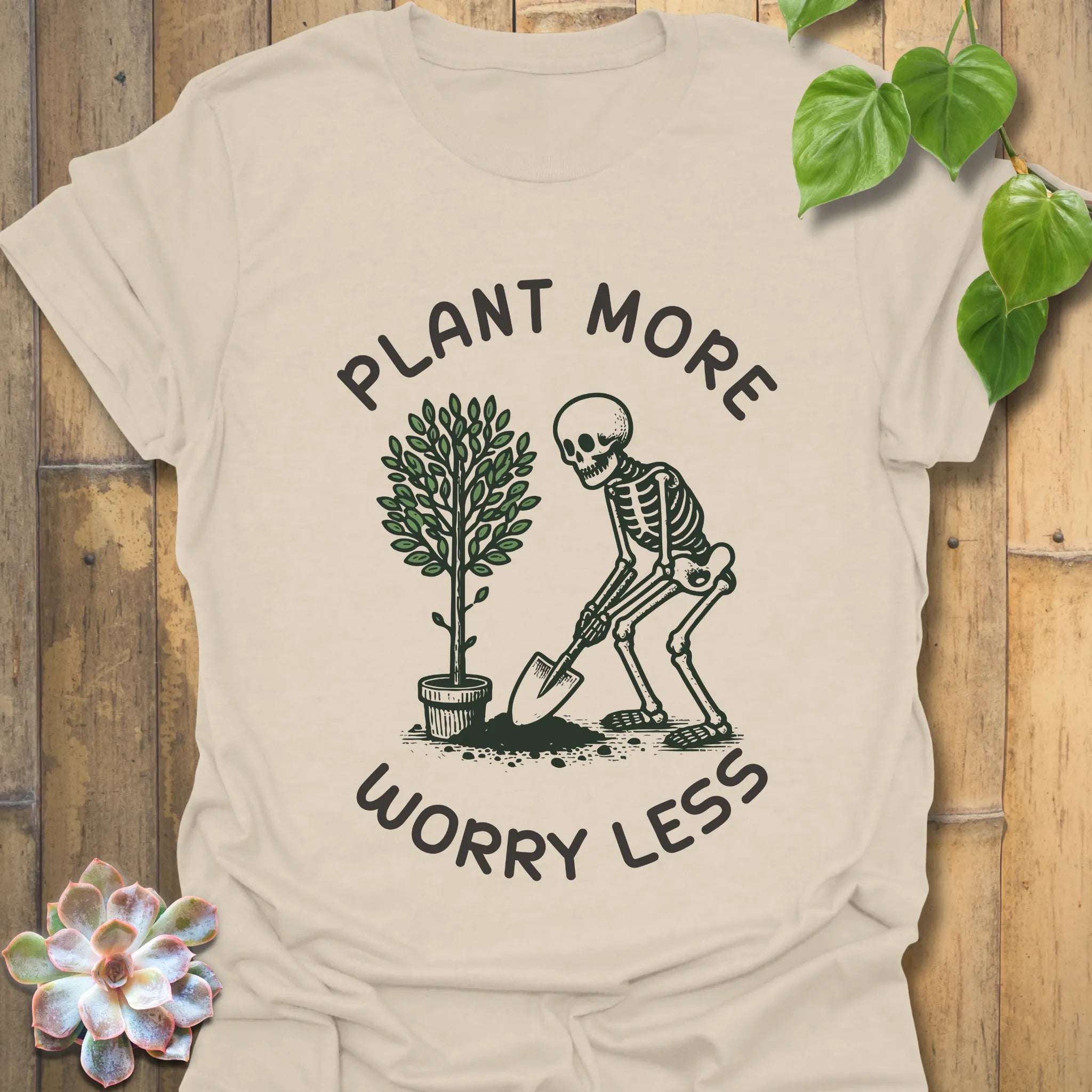 Natural colour t-shirt featuring the design Plant More Worry Less with plants and a skeleton character, sustainable casual graphic shirt.