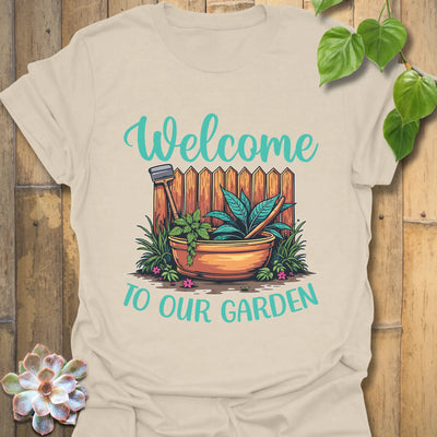 Natural colour t-shirt featuring the design Welcome to our garden with a garden scene, potted plant, and tools, making a stylish plant lover shirt and garden gift.