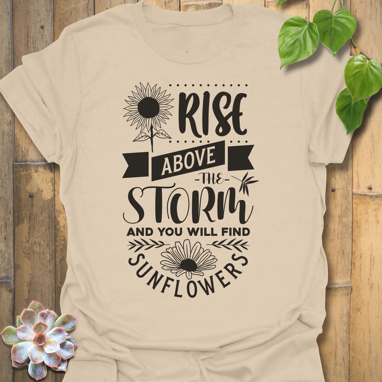 Sand t-shirt featuring the design Above The Storm printed in bold letters, floral inspirational sunflower graphic shirt.