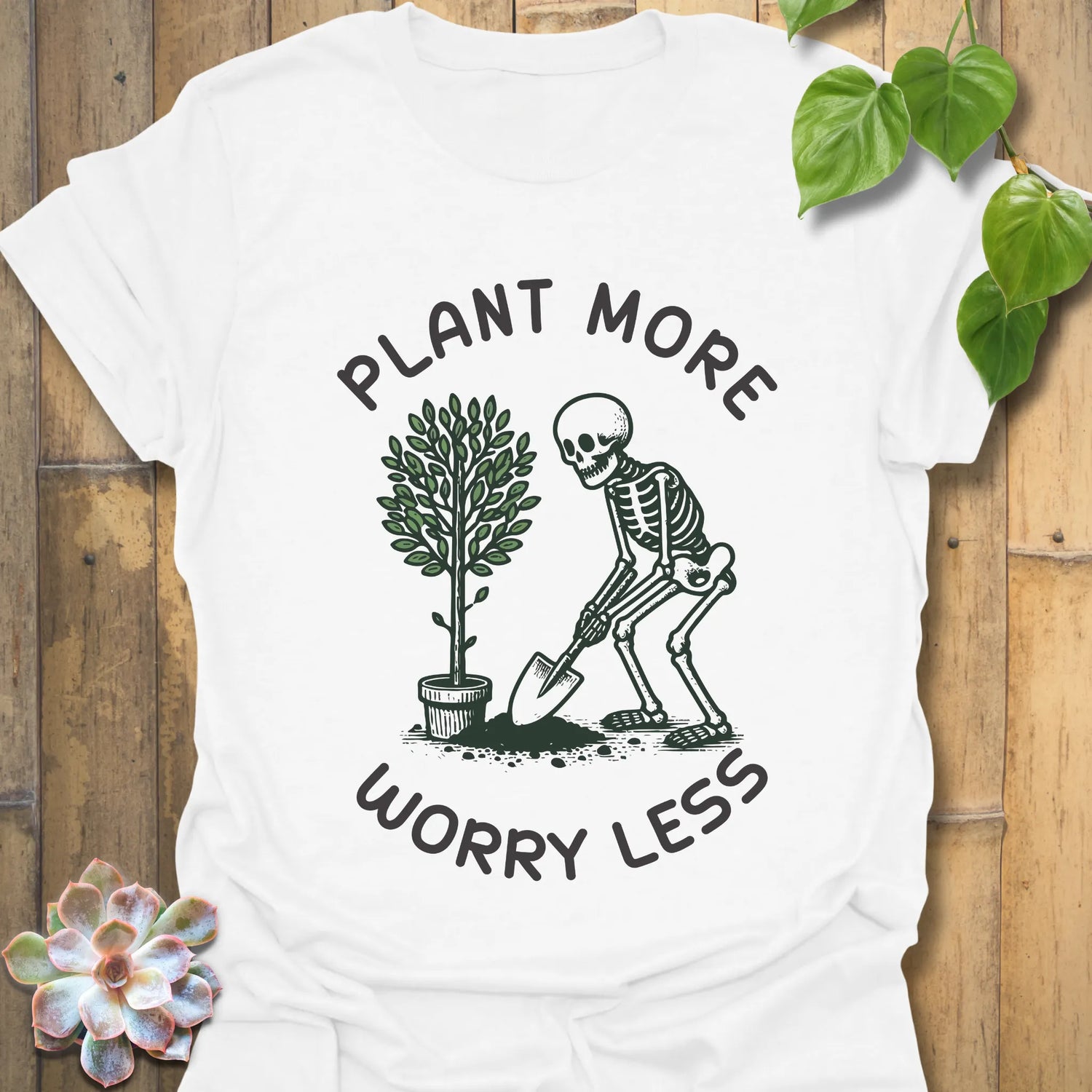 White t-shirt featuring the design Plant More Worry Less with a playful plant character and skeleton figure, eco-friendly graphic tee.