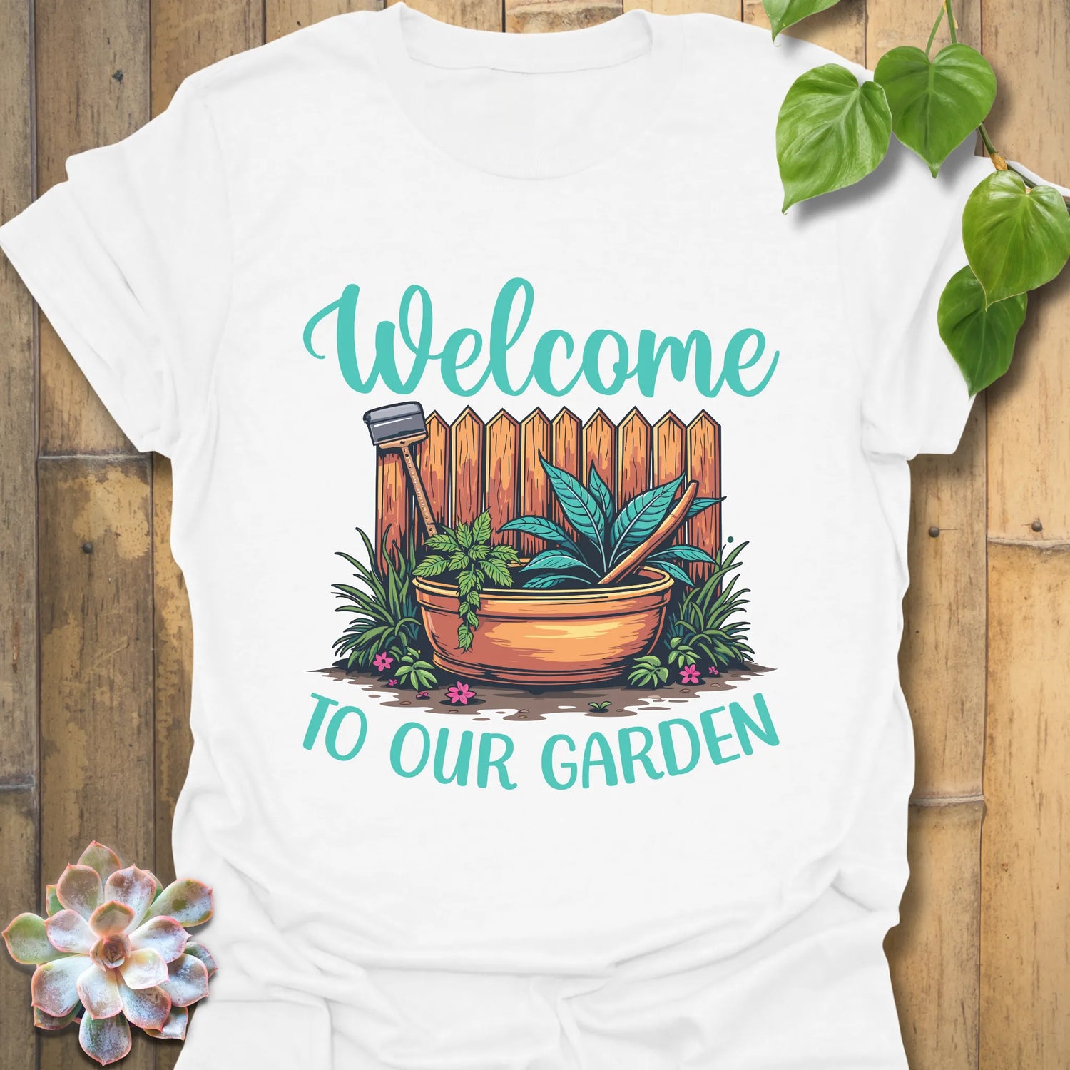 White t-shirt featuring the design Welcome to our garden with a garden scene, potted plant, and tools, perfect as a gardening graphic tee for plant lovers.