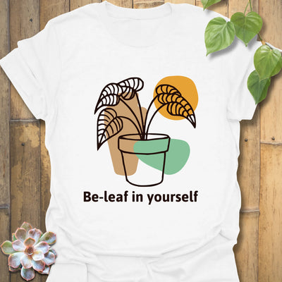 White t-shirt featuring the words “Be-leaf in yourself” along with a green plant in a pot design